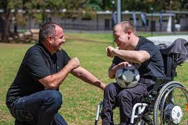 A man crouching to greet a friend who uses a wheelchair at the park